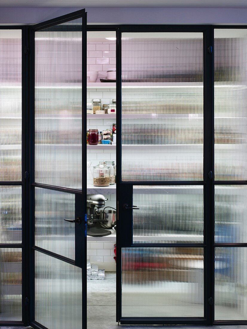 Floor-to-ceiling glass wall with black steel frame and double doors with view of open-fronted shelves in pantry