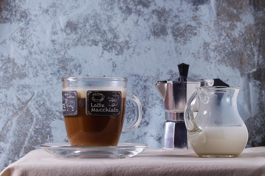 Latte Macchiato in Glastasse, Krug Milch und Espressokanne auf weißem Tischtuch