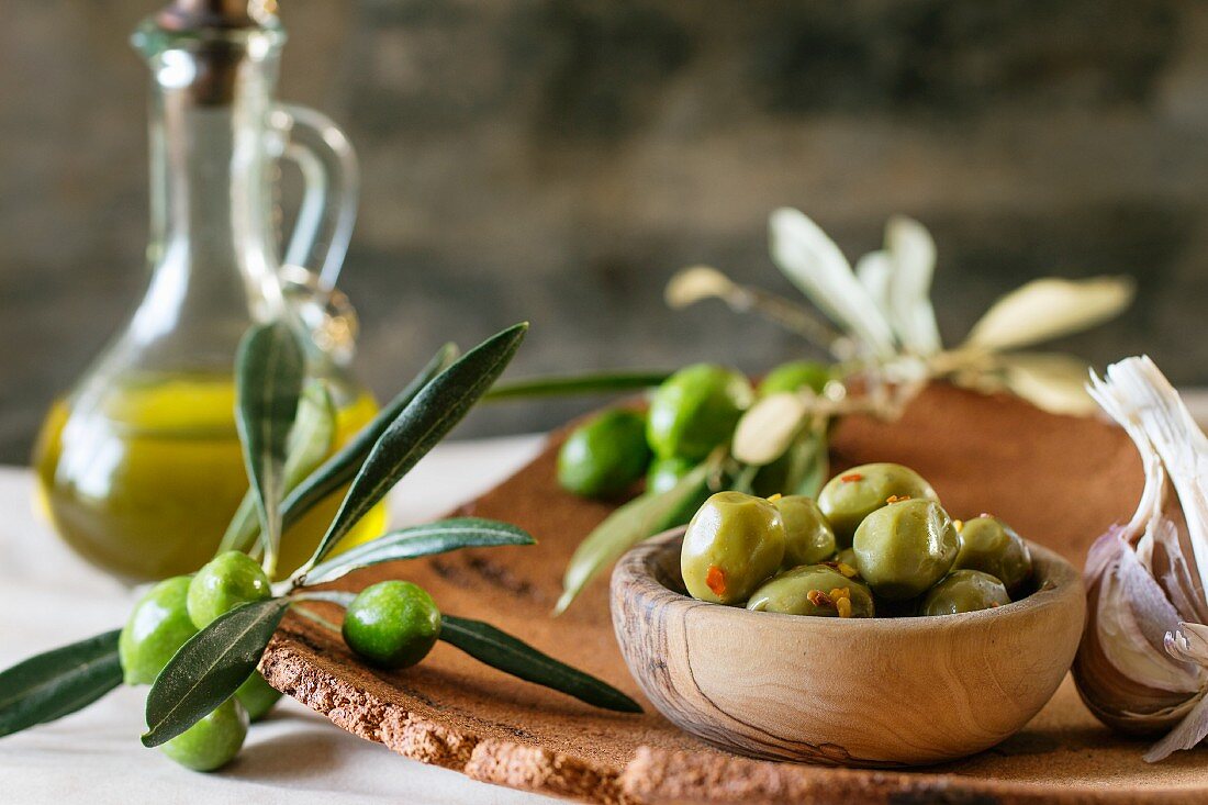Grüne marinierte Oliven in Olivenholzschüssel, serviert mit frischem Olivenzweig und Olivenöl