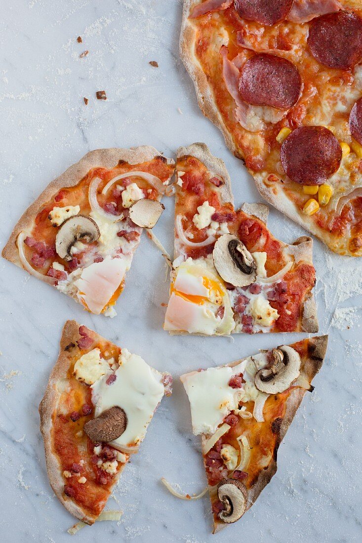 Pizza mit Tomate, Champignons, Zwiebeln, Speckwürfeln, Spiegelei (aufgeschnitten) und eine Pizza im Anschnitt mit Salami