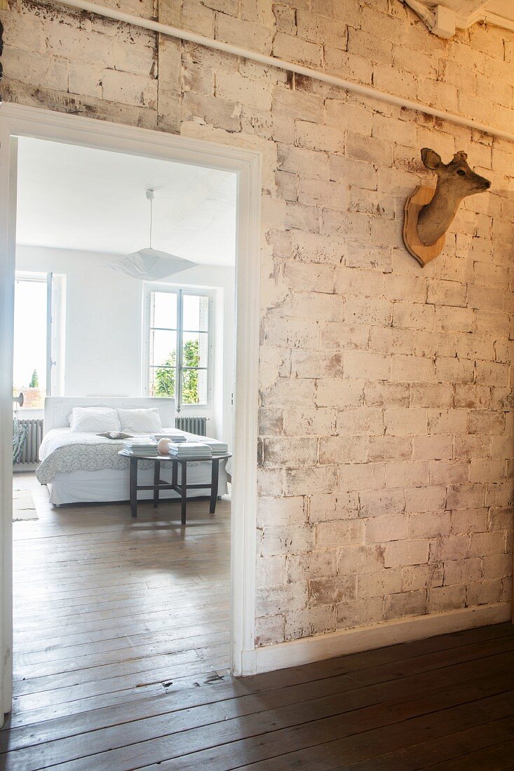 View from hallway with hunting trophy on unrendered brick wall into bright bedroom with wooden floor