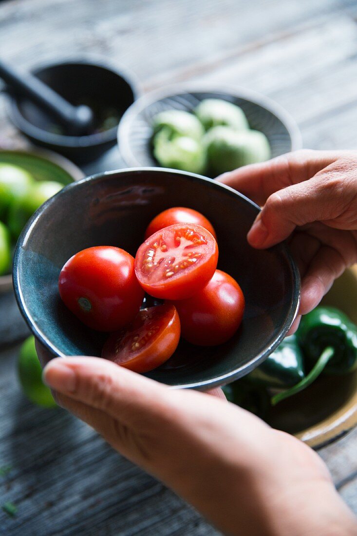 A person holding a bowl of tomatoes with tomatillos and jalapeños in the background