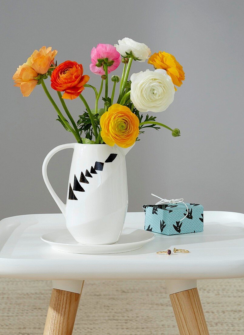 A bunch of buttercups in a white jug decorated with various tangram shapes next to a small present