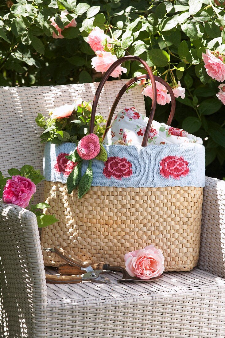 Raffia shopping bag decorated with knitted trim and knitted roses on pale rattan armchair next to climbing rose