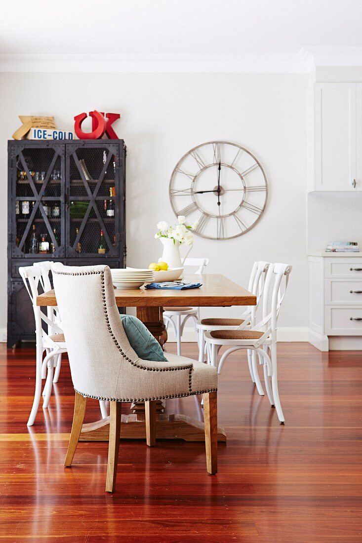 Upholstered armchairs with decorative rivets and white wooden chairs around massive dining table in front of vintage station clock and display cabinet