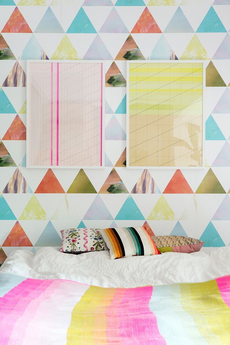 Two frame graphic artworks on colourful wallpaper above scatter cushions on bed