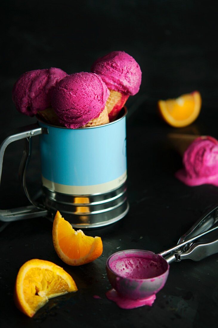 Raspberry ice cream in cones with an ice cream scoop and orange wedges