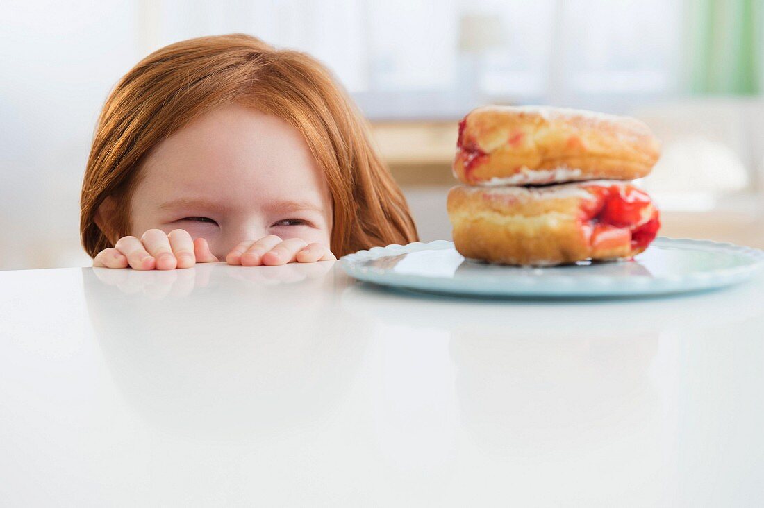The little girl looking over the edge of a table at plate of doughnuts