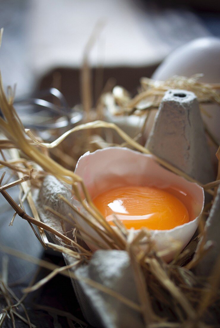A cracked egg in its eggshell in an egg box lined with straw