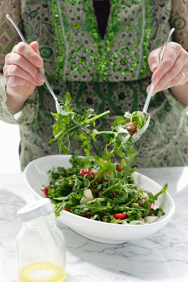 Frau mischt einen Rucola-Tomaten-Salat mit Pinienkernen und Salatsauce