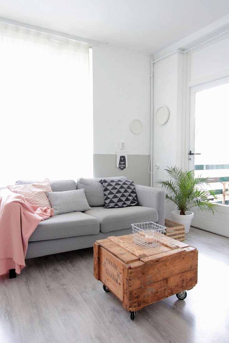 Pink blanket and various scatter cushions on pale grey couch behind vintage wooden crate used as coffee table
