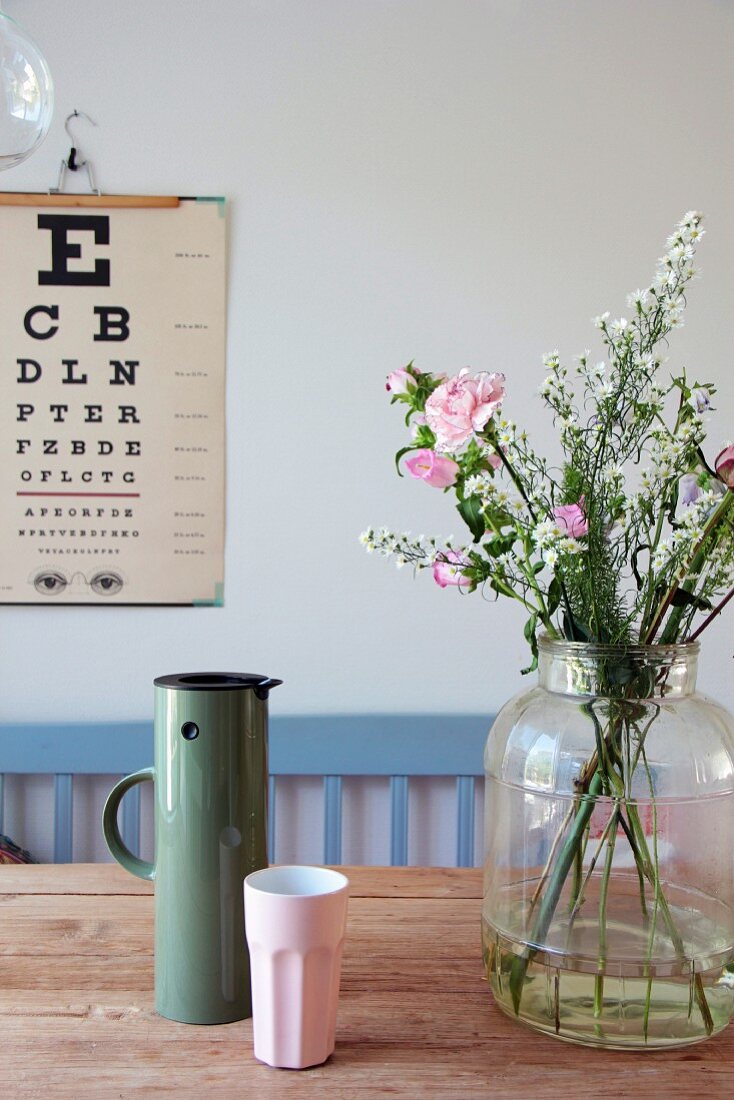 Blumen in Glasvase und Thermoskanne mit Becher auf Holztisch, Sehtafel an der Wand