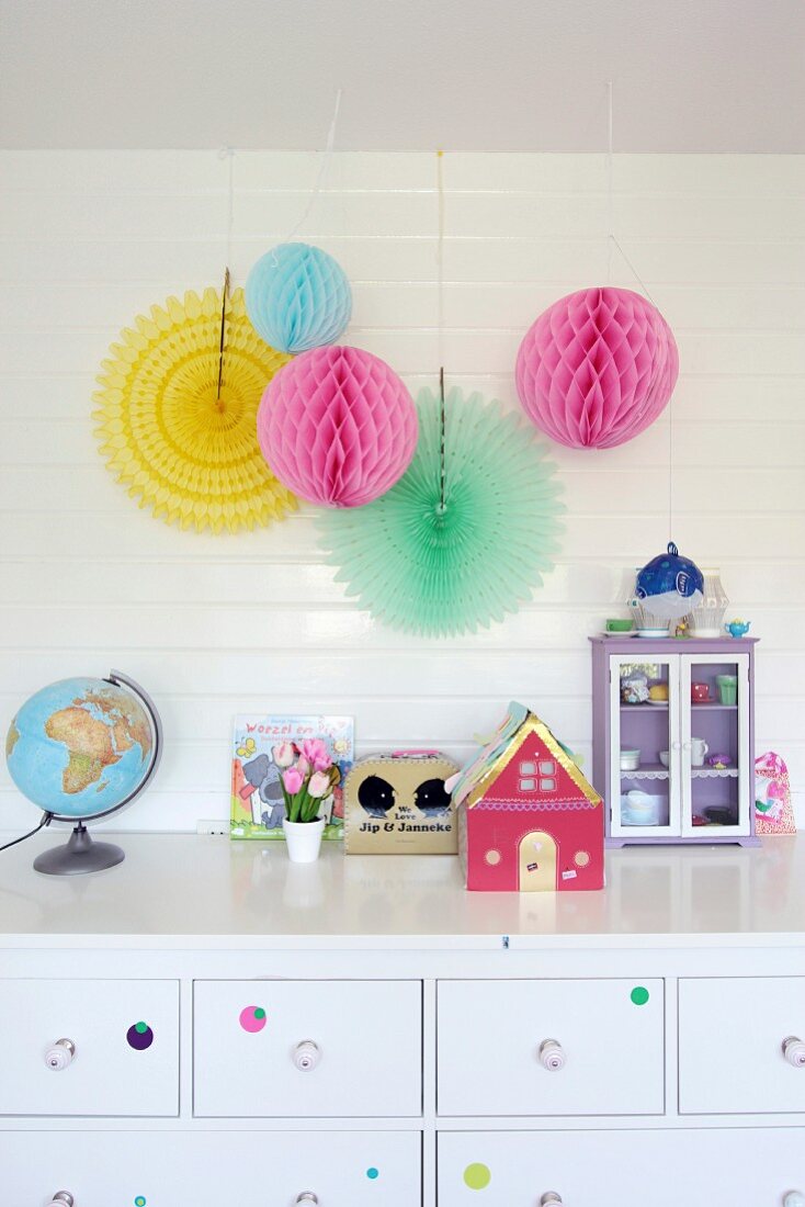 Colourful paper pompoms hung on white wood cladding above toys on chest of drawers in girl's bedroom