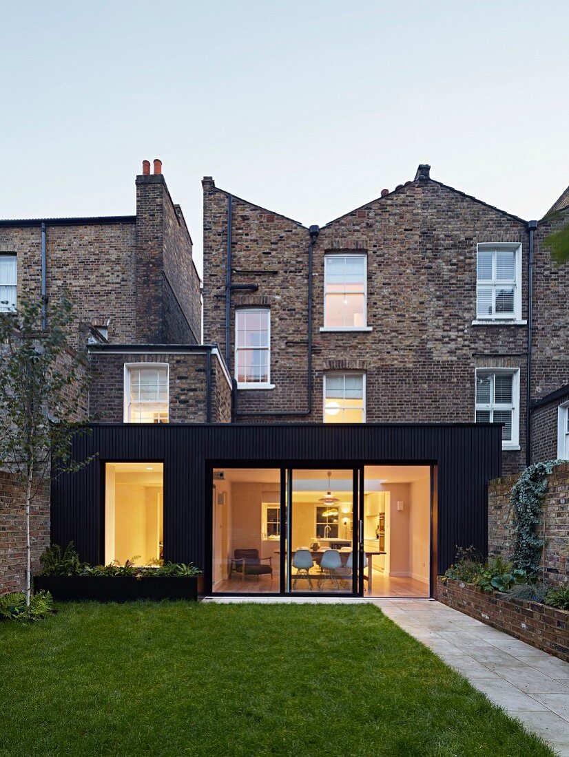 Modern extension with illuminated interior built onto traditional, English brick house seen from garden