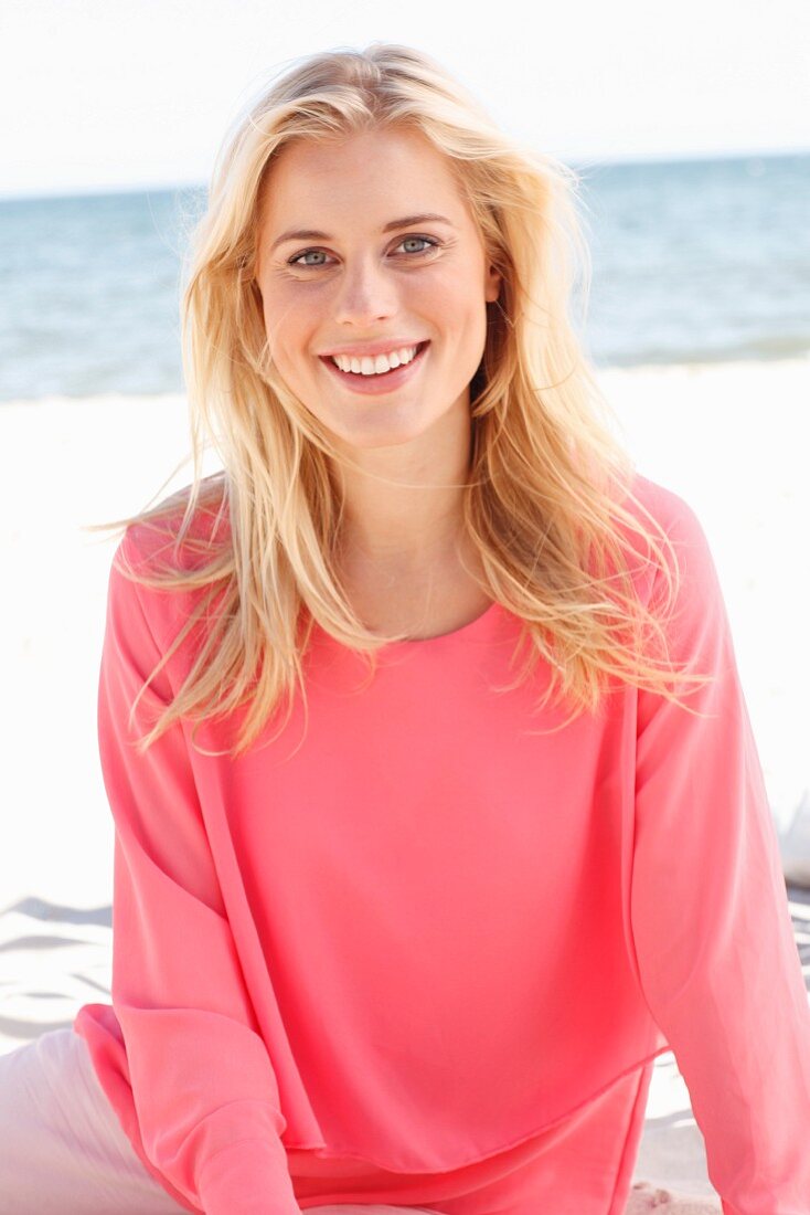 A young blonde woman sitting on a beach wearing a long-sleeved, layer-look top