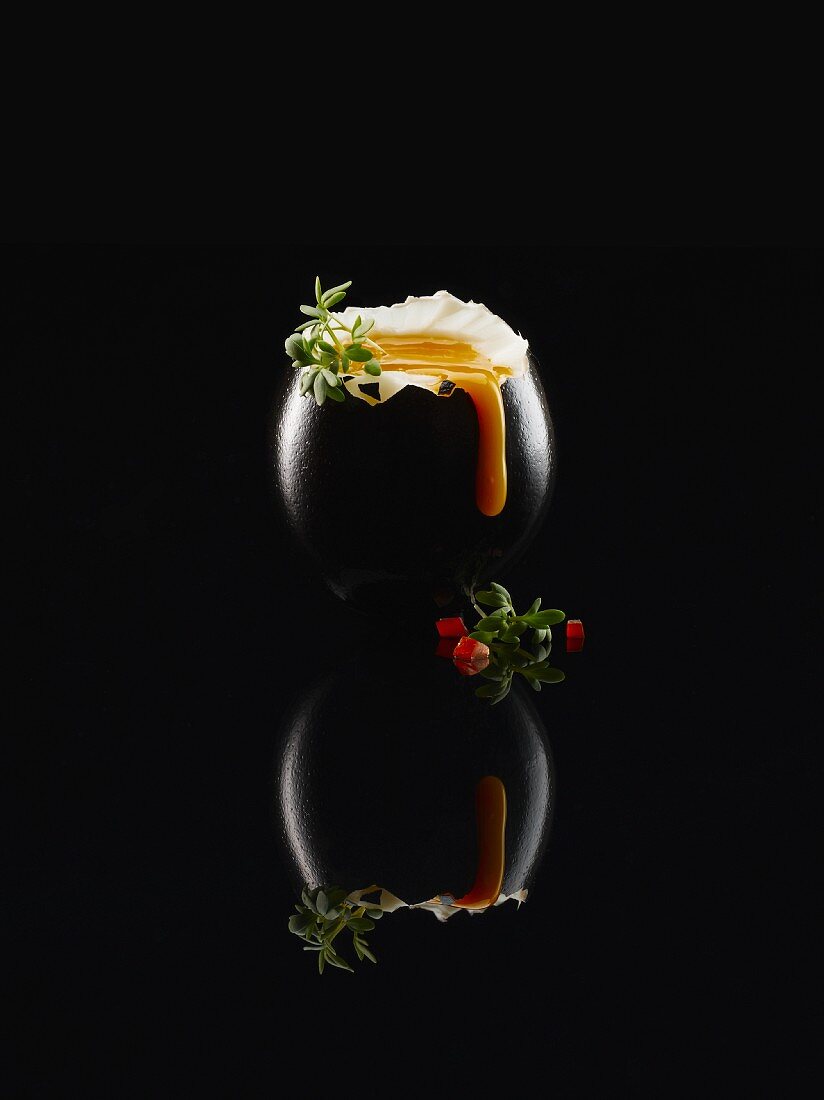 Black food: a soft boiled black egg with thyme and pieces of tomato on a black reflective surface