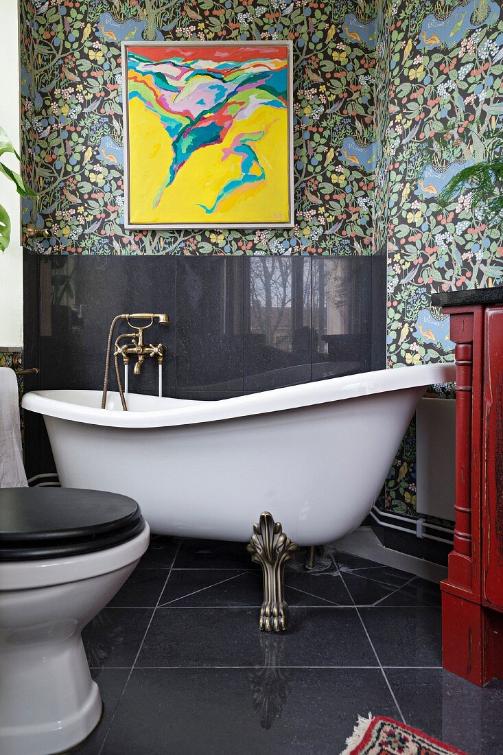 Free-standing clawfoot bathtub, black splashback on wall and modern artwork on floral wallpaper in vintage-style bathroom