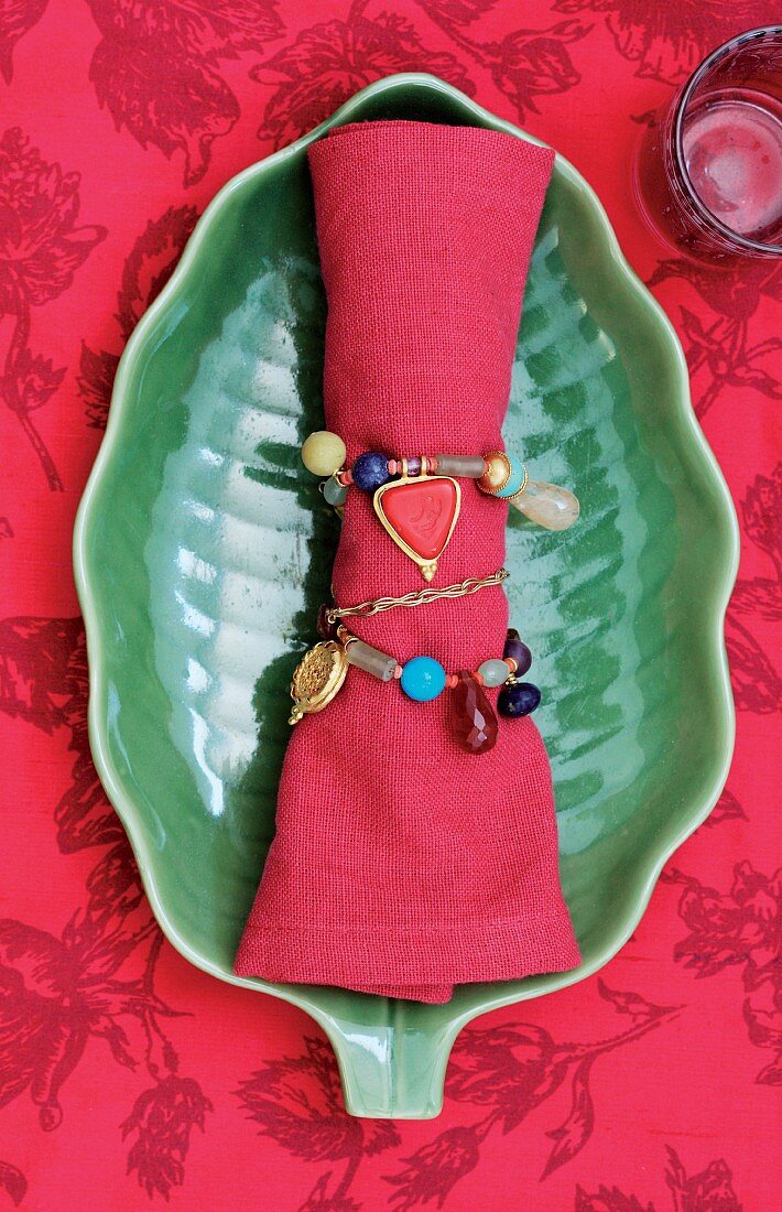 Perlenschmuck um rote Stoffserviette gewickelt auf grüner Schale in Blattform, rote Tischdecke mit Muster