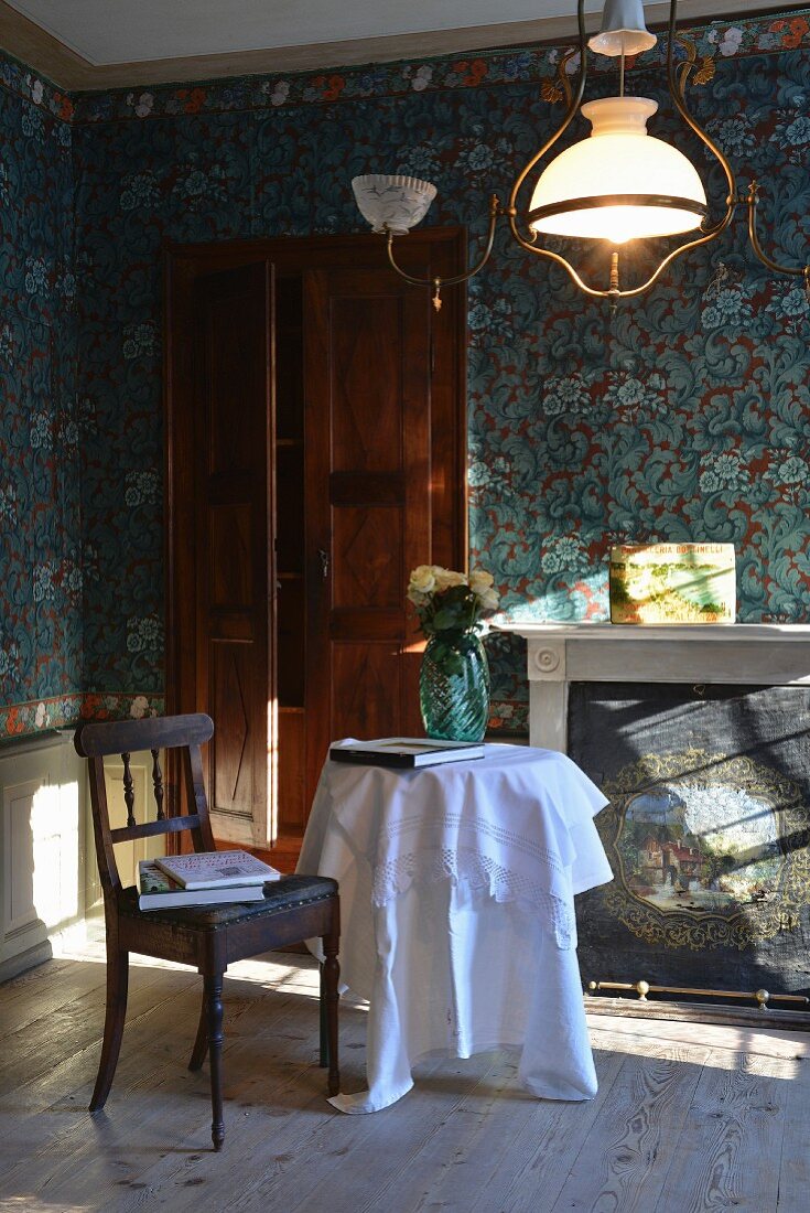 Vintage wooden chair with leather cover and round table with white tablecloth in front of fireplace in room with blue patterned wallpaper