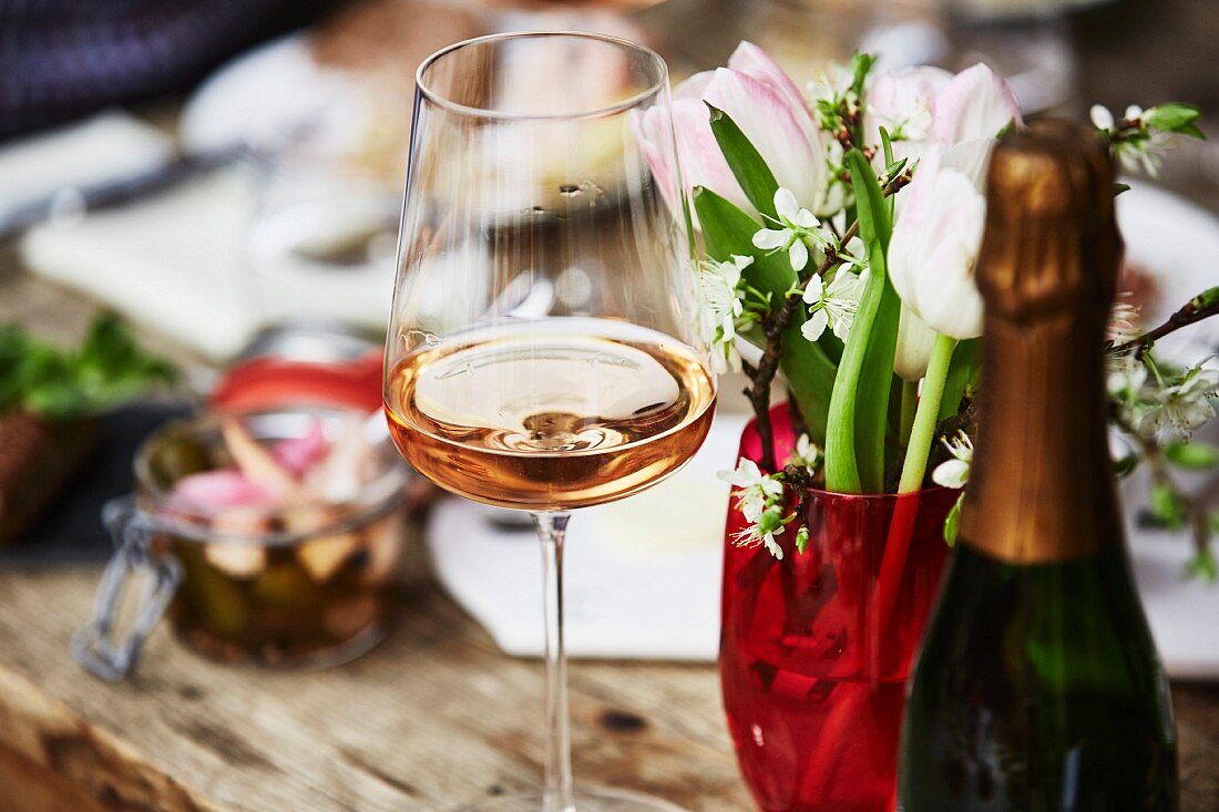 A glass of rosé champagne, a bunch of spring flowers and a bottle of champagne on a wooden table