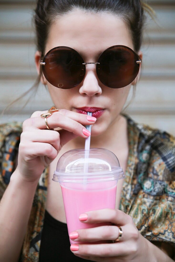 Junge Frau mit Sonnenbrille trinkt Smoothie aus Plastikbecher