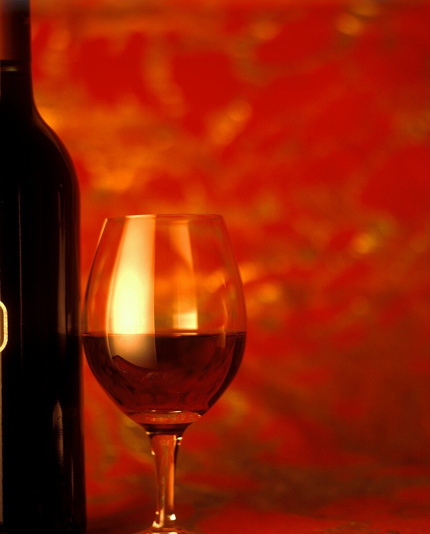 Ein gefülltes Rotweinglas neben Flasche vor rotem Hintergrund