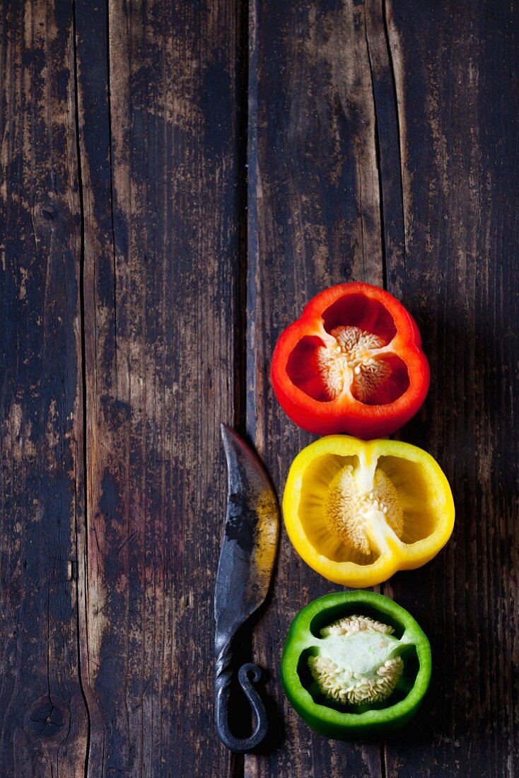 Rote, gelbe und grüne Paprika, halbiert mit Messer auf Holzuntergrund