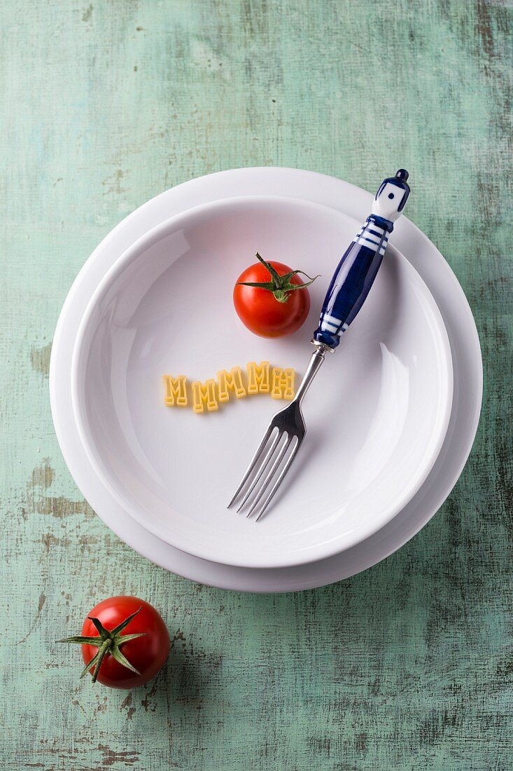 A tomato, a fork and pasta letters on a pasta plate (seen from above)