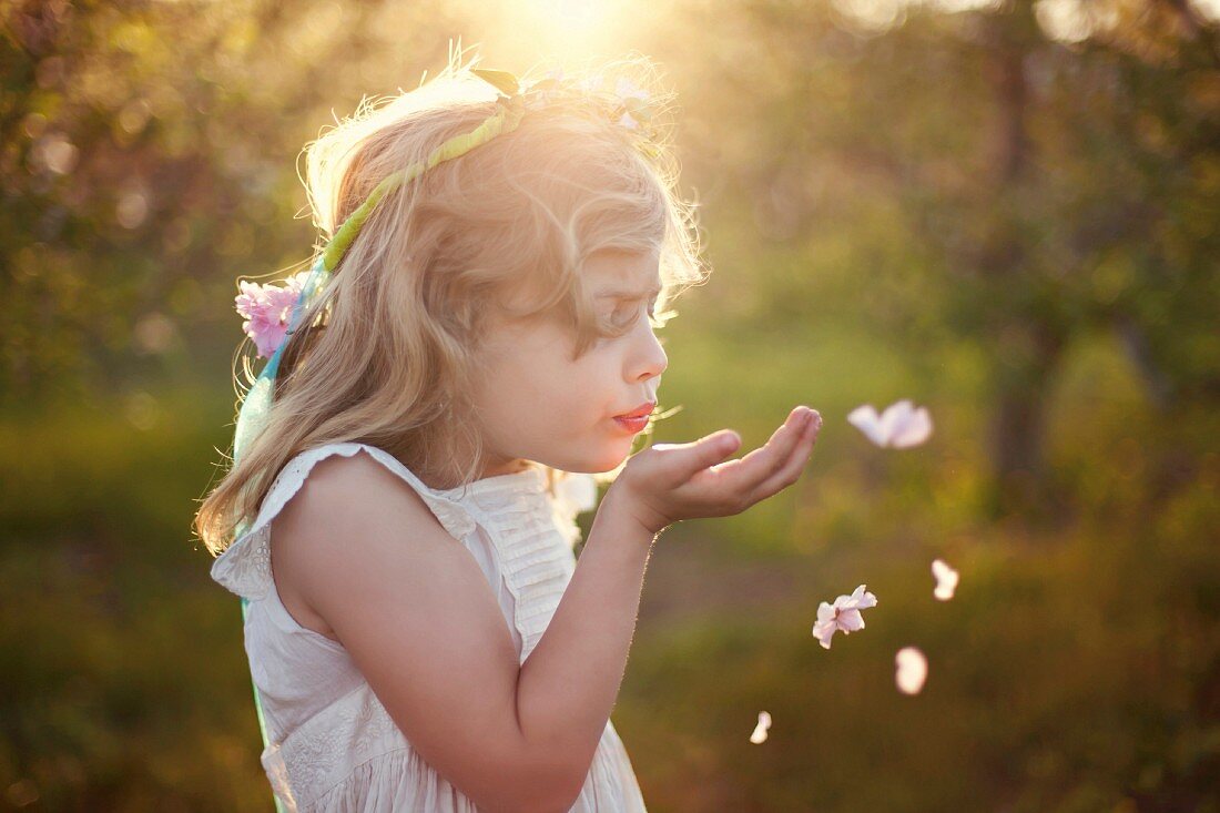 A little girl wearing a dress and a floral headband blowing flower petals away