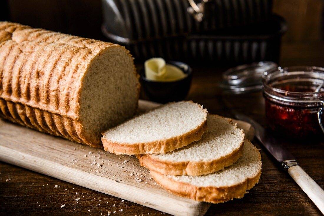 Traditional milk bread, sliced, with a baking tin, butter and a jar of jam in the background