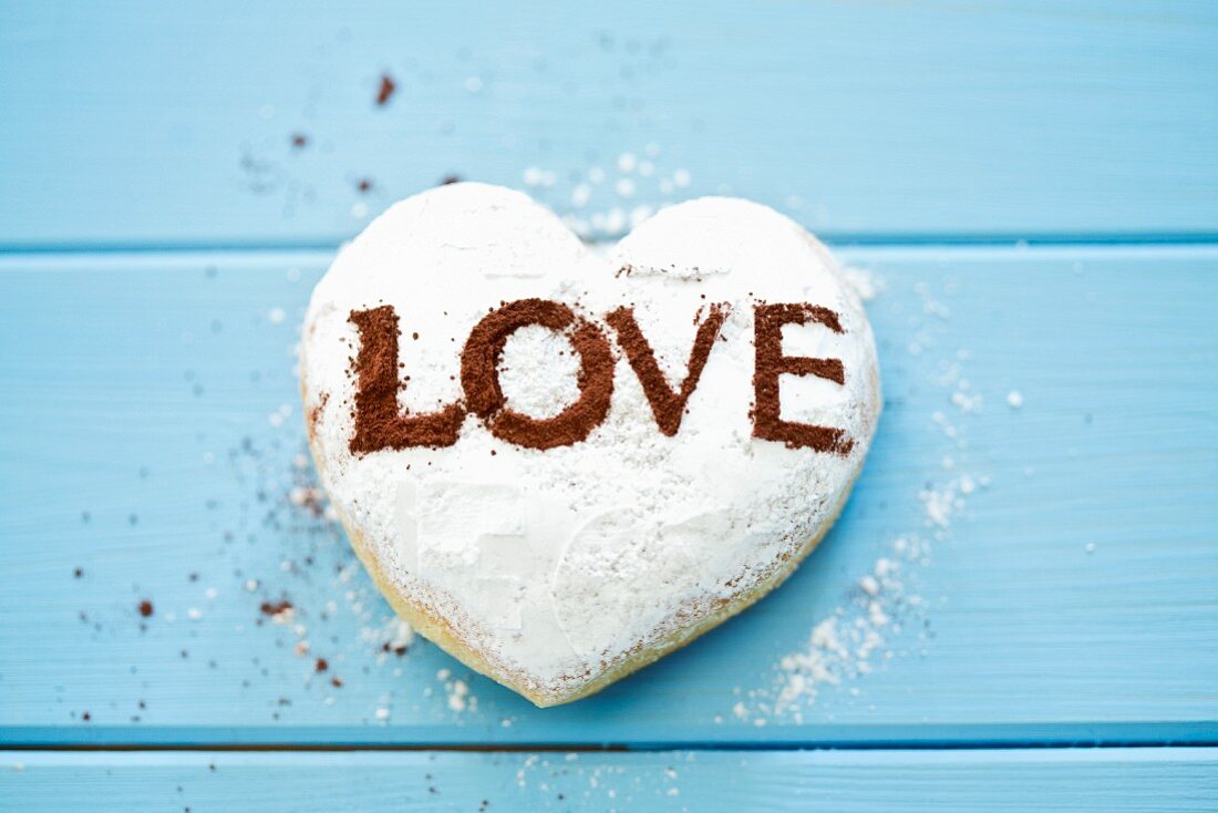 A heart-shaped doughnut with the word 'Love' in cocoa powder on the blue wooden surface
