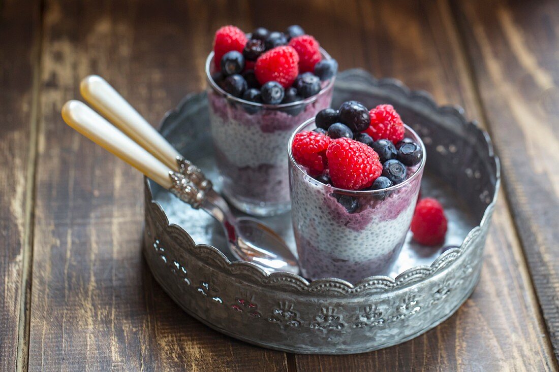 Two glasses of chia pudding with blueberries and raspberries on a metal tray