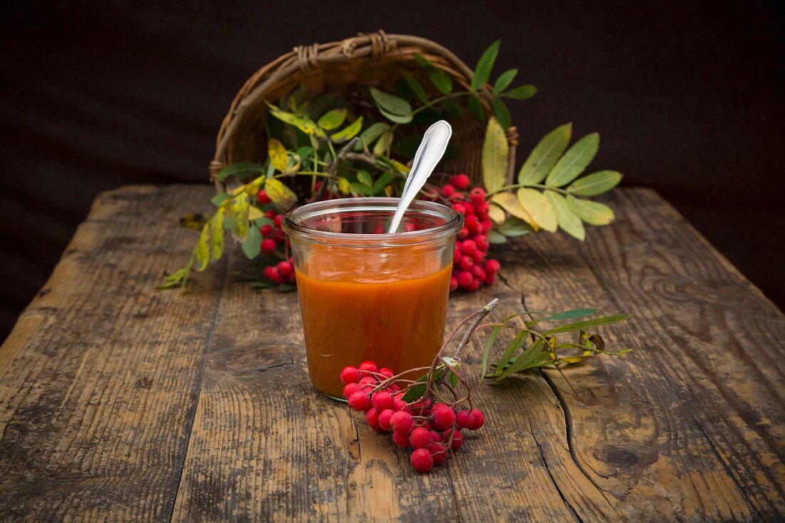 A jar of rowan berry jam with a wicker basket of rowan berries in the background