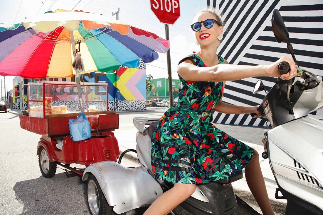 Junge blonde Frau in geblümtem Sommerkleid auf Motorroller