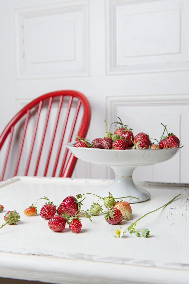 Strawberries and a porcelain bowl on a wooden table with a white wooden door and a red wooden chair in the background