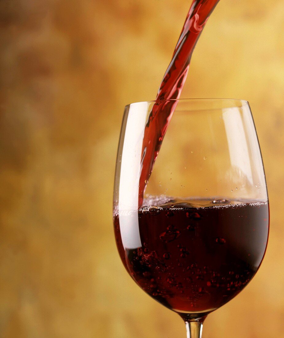 A stream of red wine flows into a red wine glass in front of a yellow background