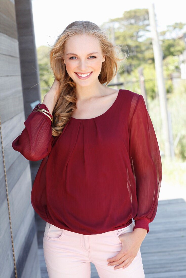 A young, dark blonde woman wearing a bordeaux red blouse and light trousers