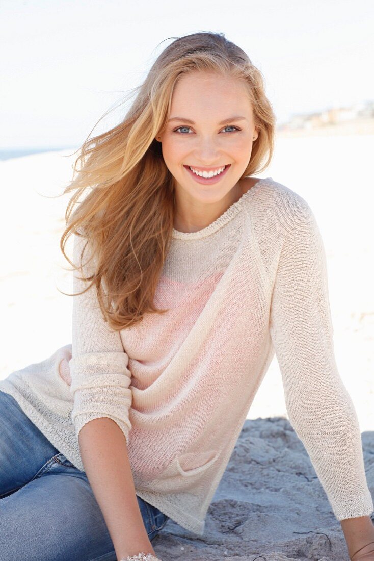 A young dark blonde woman on a beach wearing a light knitted jumper and jeans