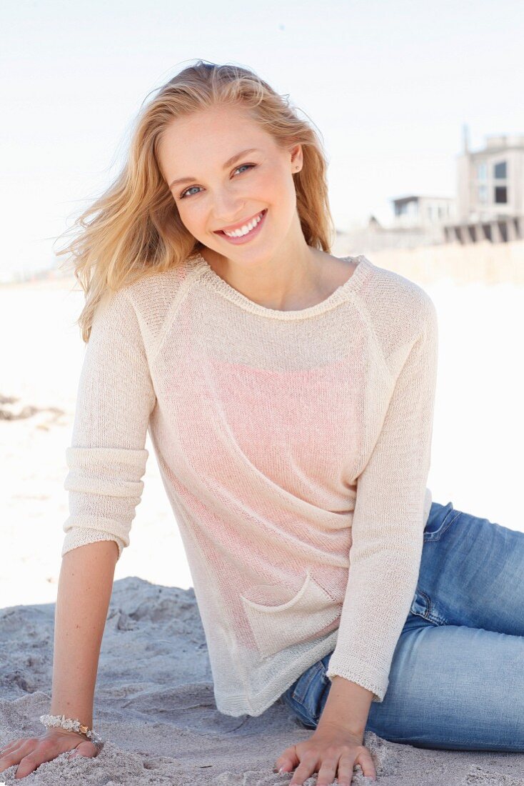 A young dark blonde woman on a beach wearing a light knitted jumper and jeans