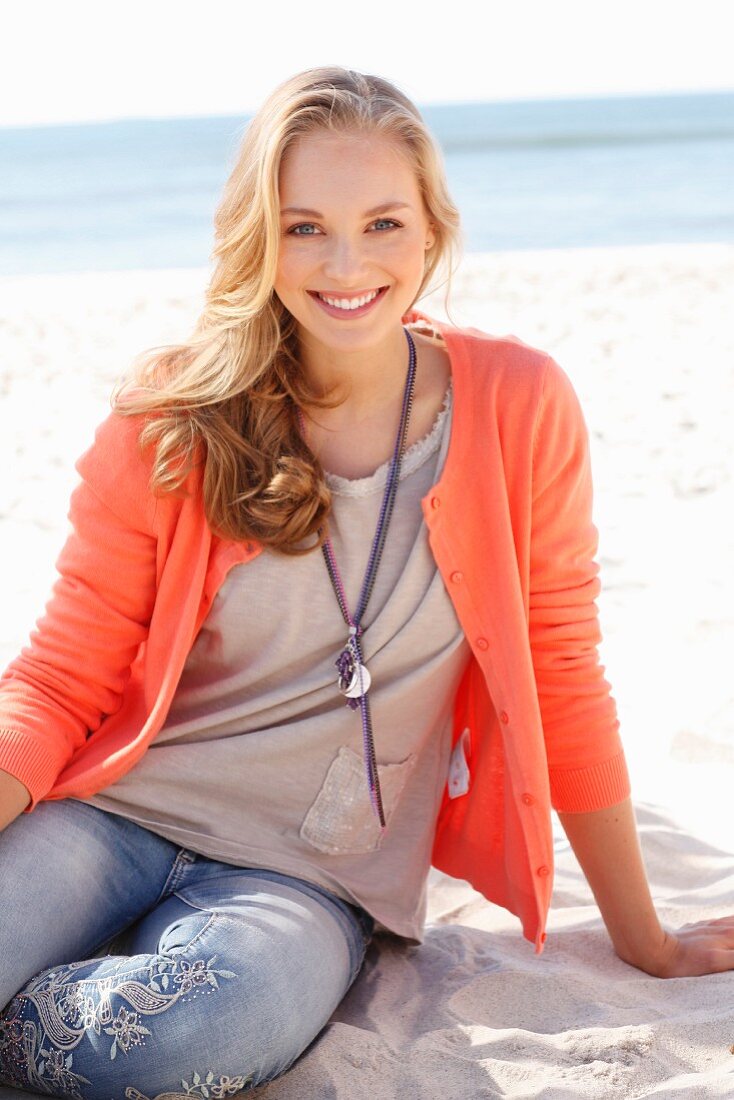 A young dark blonde woman on a beach wearing a top, a cardigan and jeans