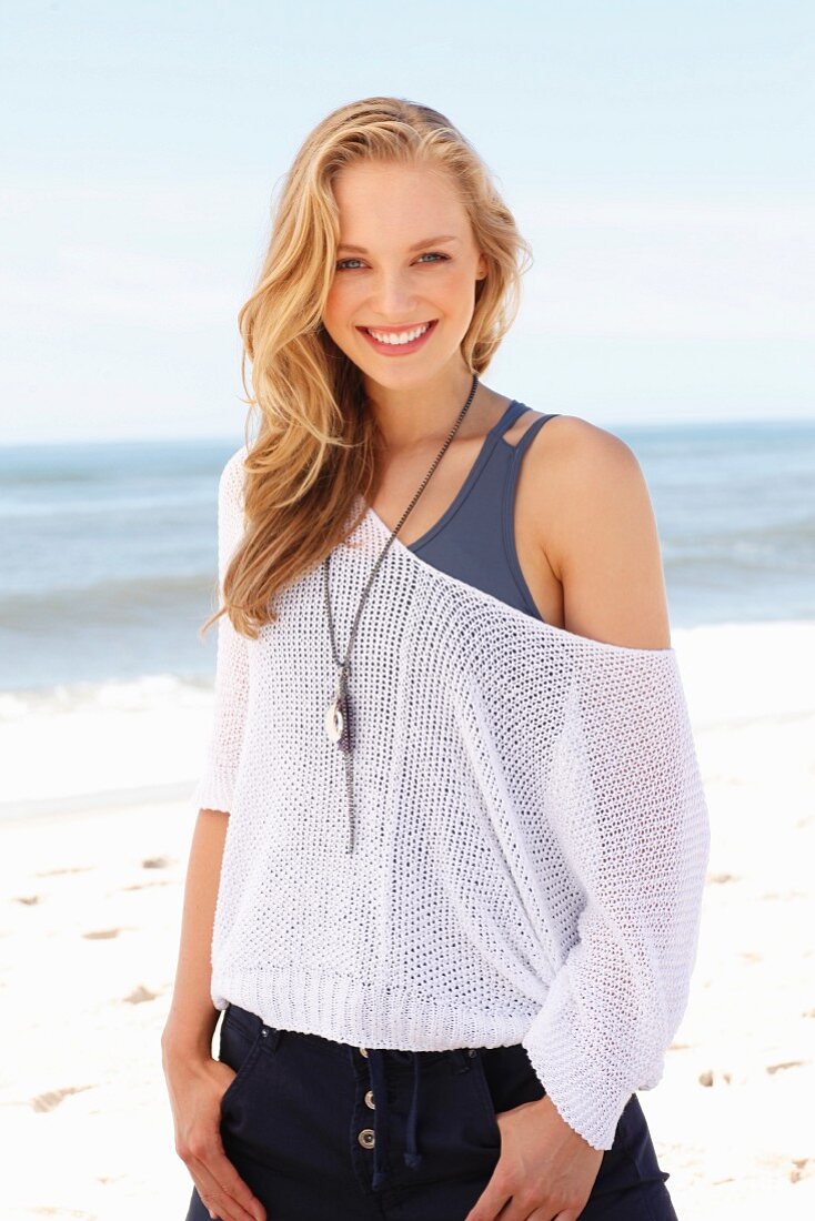 A young blonde woman on a beach wearing a tank top and a net jumper