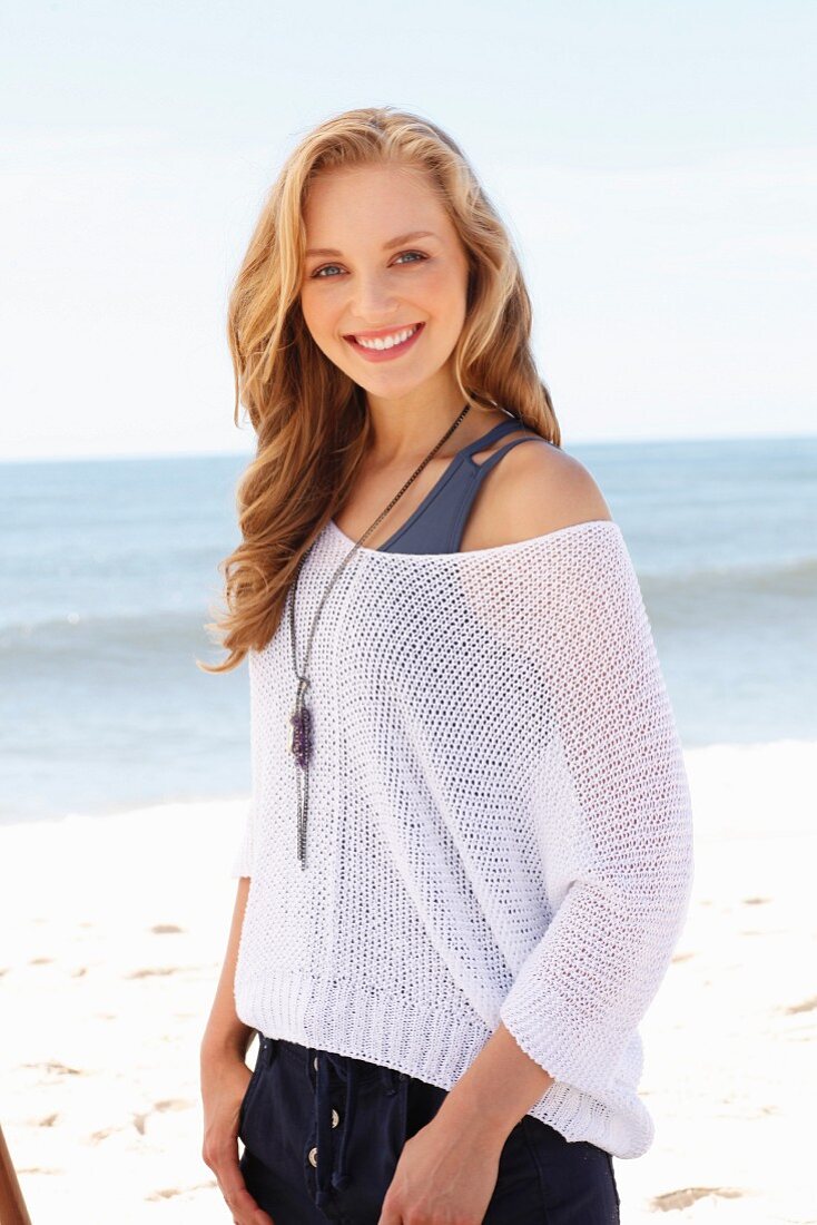 A young blonde woman on a beach wearing a tank top and a net jumper