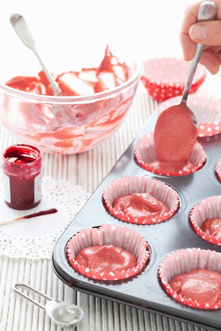 Red velvet cake batter being filled into paper cupcake cases