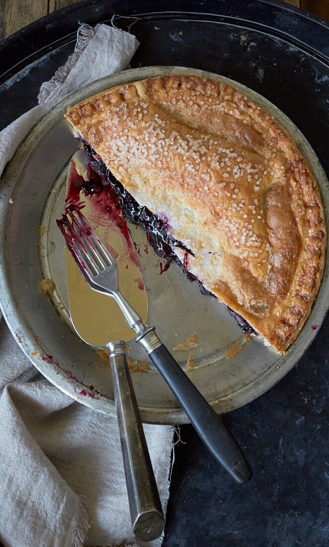 Half a blueberry pie in a baking dish with a cake slice and a fork