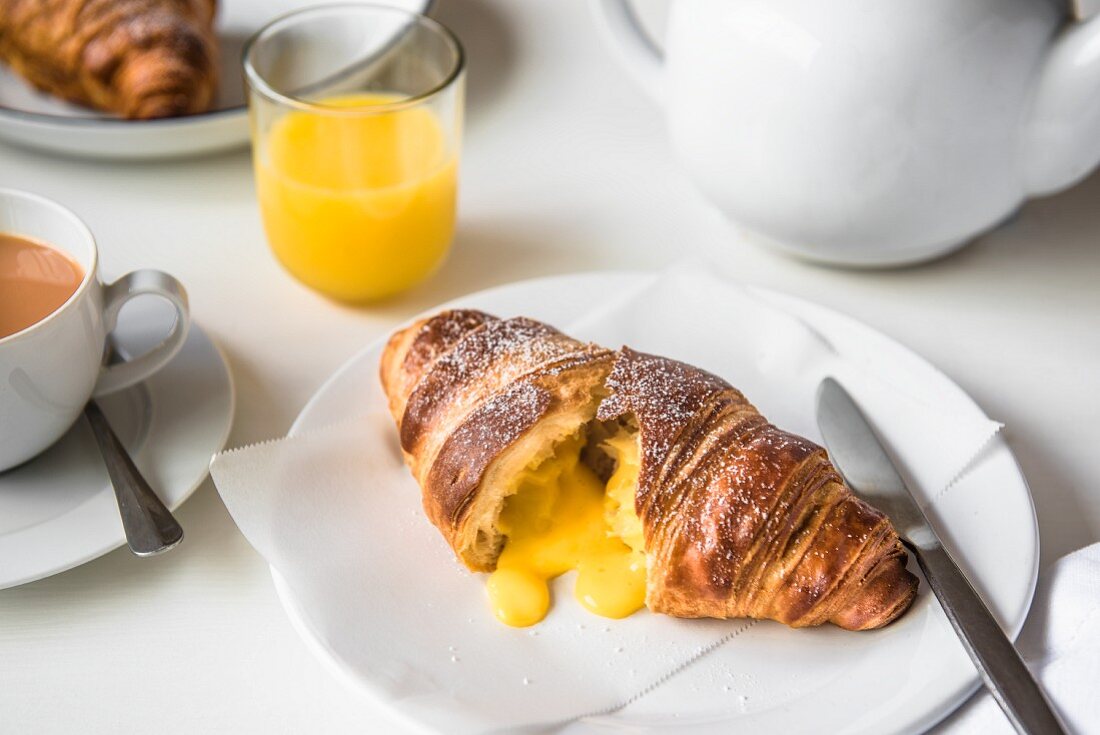 Aufgeschnittenes Croissant, gefüllt mit gesalzenem Eigelb, auf Teller mit Tee und Orangensaft