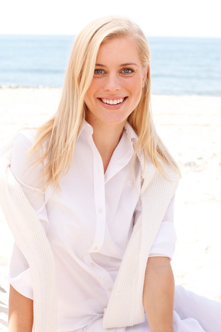 Junge blonde Frau in weisser Bluse und Hose sitzt am Strand