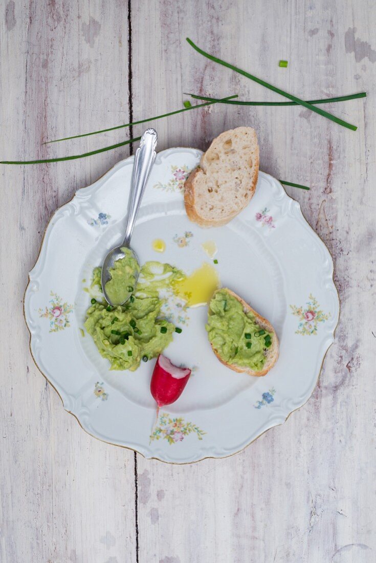 Avocado cream with chives and olive oil on a porcelain plate with a slice of baguette and radishes