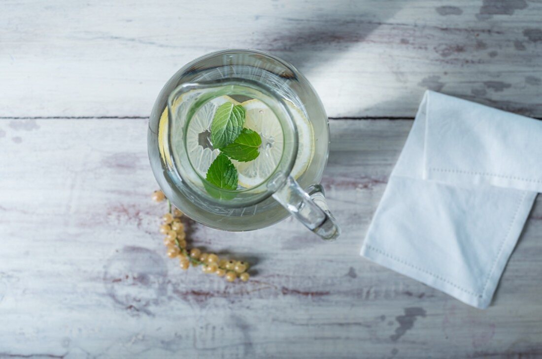 A glass water jug with lemon and mint on a white wooden surface next to whitecurrants and a fabric napkin