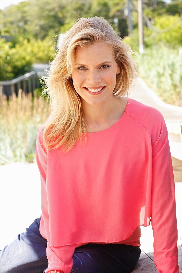 A blonde woman wearing a long-sleeved, layered-style, salmon-coloured shirt and blue trousers