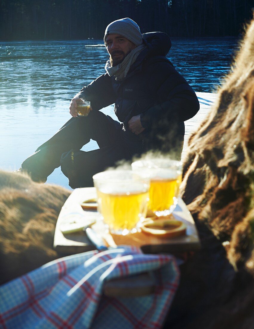 Warm angezogener Mann sitzt mit heißem Apfelcidre an Gewässer